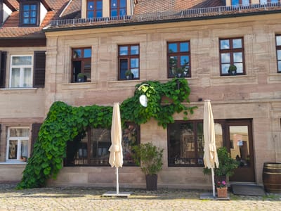 The image shows a facade of a historic building in Erlangen, Germany. The building has stone walls, brown-framed windows with multiple panes, and a tiled roof. Lush green ivy covers a significant portion of the lower facade, cascading from a window area and arching over a doorway. Two closed cream-colored patio umbrellas stand on the cobblestone ground in front of the building, flanking a potted green plant. To the right of the umbrellas and plant, there is a wooden door and a large wooden barrel. The scene appears to be an outdoor seating area for a cafe or restaurant. The windows reflect the sky and surrounding environment.