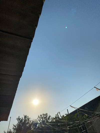 The image shows a bright, clear sky with the sun low on the horizon, casting a soft glow. In the foreground, a dark, textured overhang juts into the frame from the left. Below the sun, the tops of trees are visible, their leaves dark against the light. In the lower right, the angled roof of a building is partially obscured by what appears to be a string of lights or decorations. A utility wire stretches across the scene, with small pieces of white material attached. A faint, small, circular green light is visible higher in the sky. The scene suggests an outdoor setting, possibly a backyard or rooftop in Babayurt, Russia, during late afternoon or early evening. There are no people or animals visible.