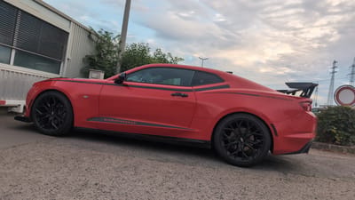 A bright red Chevrolet Camaro coupe with black racing stripes and a large rear spoiler is parked on a gravel surface in front of a corrugated metal building. The building has a horizontal window with dark louvers. Green bushes and trees are visible behind the car and to the right. In the background, there are tall power line towers and a circular traffic sign. The sky is cloudy. The scene is located in Worms, Germany. There are no people or animals present.