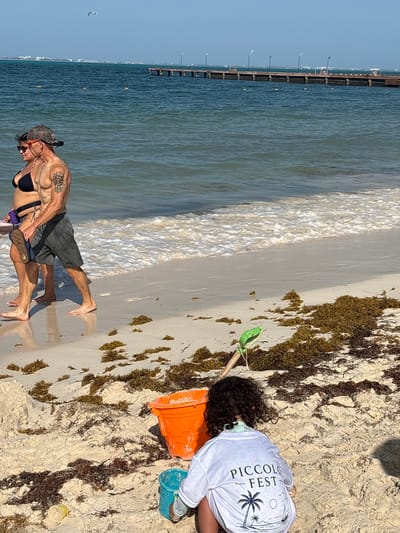 At a beach in Punta Sam, Mexico, a couple walks along the shoreline. The man, wearing a camouflage baseball cap, sunglasses, and dark shorts, has a muscular build and a tattoo on his upper left arm. The woman, in a black bikini and sunglasses, carries a flip-flop in her right hand. They are walking towards the left side of the frame.

In the foreground, a child with dark, curly hair is crouched down on the sand. They are wearing a white long-sleeved shirt with the text "PICCOLO FEST" and a graphic of palm trees and a sailboat. The child is near a bright orange bucket, a smaller turquoise bucket, and a green shovel, suggesting they are playing or building in the sand.

The beach is covered in sand and scattered with brown seaweed. The ocean water is a clear blue-green, with waves gently breaking on the shore. In the background, a long wooden pier extends out into the water, with lampposts lining its length. The sky is clear and blue.