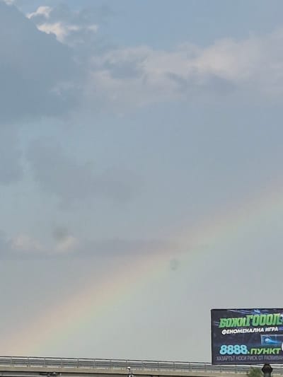 The image shows a cloudy sky with a faint rainbow visible on the right side. A billboard with text is also visible on the right. The sky appears to be a light blue or gray, indicating a potentially overcast or post-rainy day. The overall scene suggests an outdoor setting.

The billboard displays text in a vertical orientation. The prominent text is in green with a white outline and reads "БОЖИ ГОООЛЕ". Below this, in smaller white text, it says "ФЕНОМЕНАЛНА ИГРА". Further down, in a different font and color, are the numbers "8888.ПУНКТ". Beneath this, in smaller white text, is "ХАЗАРТЪ НОСИ РИСК ОТ РАЗЕИВА".

The time of day is not explicitly stated, but the lighting suggests it could be daytime. The weather appears to be cloudy with a possibility of recent rain, given the presence of the rainbow. The location cues from the provided context are Krustina, Bulgaria. No people are clearly visible in the image, and the focus is on the sky and the billboard. The overall emotion conveyed is neutral, with a slight sense of atmosphere due to the rainbow and clouds.