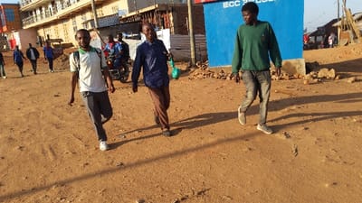 The scene takes place in the city of Kayna, Democratic Republic of the Congo, at latitude -0.561 and longitude 29.179. Four men are walking along a dirt road. The man in the front is wearing a green shirt, blue jeans, and white sneakers. The man in the middle is wearing a blue button-down shirt, brown pants, and black shoes. The man behind him is wearing a white shirt, gray pants, and white sneakers. The man in the back is wearing a light blue shirt, jeans, and white sneakers. All the men are walking in the same direction. The dirt road is lined with a few buildings, and a blue wall with the letters "ECC" on it is visible on the right side of the frame. The ground is covered with dirt and sand, and there are some rocks scattered around. The sky is blue, and the sun is shining.