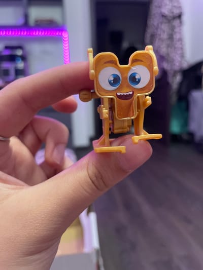 A close-up, eye-level shot shows a person's hand holding a small, orange, toy robot character. The robot has large blue eyes, a wide open mouth with white teeth, and appears to be made of plastic. The robot is held between the thumb and index finger, with other fingers visible below. In the background, out of focus, is a dimly lit room with what appears to be purple LED strip lighting and some clothing hanging. There is no indication that this scene is in Borisova Griva, Russia.