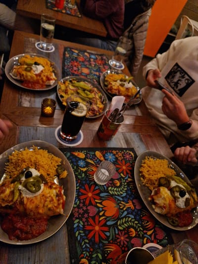 The image depicts an indoor dining scene at a restaurant in Aschaffenburg, Germany, characterized by warm, dim lighting. A rustic wooden table with a distressed panelized surface is prominently featured, covered with colorful placemats displaying elaborate floral and leaf patterns in red, orange, blue, and green hues.
Several individuals are gathered around the table, though only partially visible. On the right, a person wearing a white hoodie with a black square graphic is holding a smartphone with both hands, appearing to interact with it or take a photo. Another person's arm is visible on the left side of the table. A third individual in a dark garment is partially visible in the upper left background.
The table is laden with multiple dishes of food and beverages. In the foreground, two plates each contain a large, cheese-covered burrito or enchilada, topped with sour cream, sliced green jalapeños, and a black olive, accompanied by a generous portion of yellow rice and a side of red salsa. Towards the center, a tall glass of a dark beverage with a lemon slice rests on a coaster. Adjacent to it is a small, lit tea light candle within a golden, perforated holder. Another plate further up contains what appears to be nachos or a similar dish with melted cheese, sliced green peppers, and red onions. In the upper left, a glass of water with a lime slice is visible on a coaster, next to another plate of food resembling the enchiladas. A red metal container filled with forks and knives stands in the middle of the table. A transparent empty glass is also present. An orange wall is visible in the background on the right.