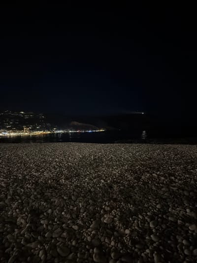 This is a nighttime photograph taken from a pebble beach in Xàbia/Jávea, Spain. The foreground is dominated by a vast expanse of light-colored pebbles, unevenly distributed and casting shadows in the low light. In the mid-ground, the dark, calm sea stretches towards the coastline. The background reveals the illuminated town of Xàbia, with clusters of buildings and streetlights dotting the hillside and lining the waterfront. A faint light, possibly from a boat or a distant lighthouse, is visible on the horizon over the sea. The sky is a deep, dark blue, characteristic of a clear night.