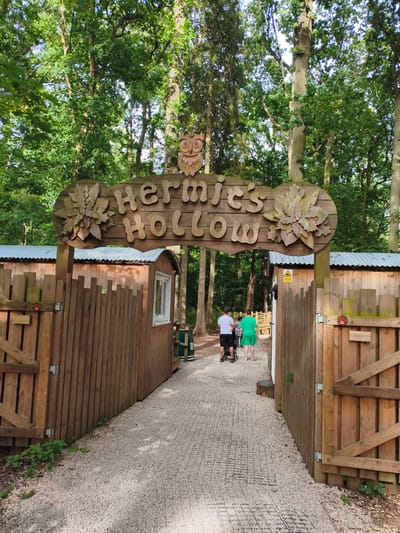 The image shows a wooden archway with the words "Hermits Hollow" carved into it, topped with an owl carving. The archway leads to a gravel path lined with wooden fences. Two people, one wearing a white t-shirt and black shorts, and another wearing a green dress, are walking away from the camera down the path, with a stroller between them. Behind the archway, there are wooden structures with corrugated metal roofs. The scene is set in a wooded area with tall trees. This is a scene at The Glebe, a country park and leisure site located in Rugby, United Kingdom.