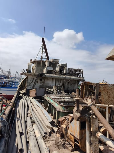 This image captures a disused and heavily rusted ship, likely a fishing vessel, in a port or shipyard setting in Chimbote, Peru. The ship's superstructure is heavily weathered, with peeling paint and exposed metal. Various pipes, some large and cylindrical, are piled on the deck, suggesting they are either part of the ship's systems or debris. In the background, other vessels, also appearing to be fishing boats with numerous poles and rigging, are visible.
The scene is set outdoors under a partly cloudy sky with bright blue patches. The sun appears to be high in the sky, indicating it is likely daytime. The overall impression is one of decay and abandonment, with the rusty metal and disheveled state of the ship conveying a sense of neglect. The setting clearly points to a maritime environment.
A notable detail is the visible text "CATERPILLAR" on a yellow piece of equipment in the foreground, likely a generator or engine, further reinforcing the industrial and maritime context. The composition focuses on the details of the decaying ship, highlighting its structural elements and the accumulated detritus.
