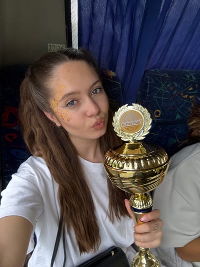 A young woman poses with a gold trophy in what appears to be the interior of a bus. The woman is lying down, with the trophy held above her. She has glitter on her face. The trophy has an emblem on it that reads "Artist Hub Dance Competition 2026". The bus has blue seats. The number "2726" is visible on a panel. The location is not apparent from this image and a specific location in Strumica, North Macedonia cannot be determined.