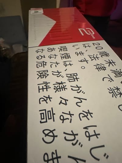 The image shows a white and red cigarette carton from the brand "2 CARTONS". The carton has Japanese writing on its side with the message, "Smoking causes lung cancer."