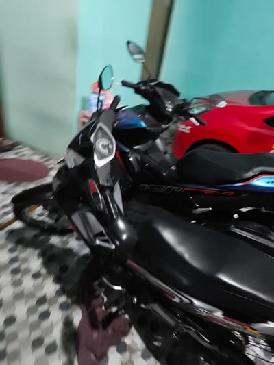 A black motorcycle is parked in a room with light blue walls and a patterned floor. A red car is visible in the background behind the motorcycle.
