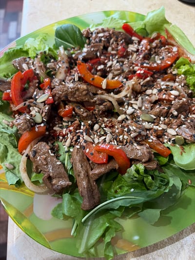 A close-up, overhead shot shows a colorful salad on a green and yellow patterned plate. The salad features sliced beef, red bell pepper strips, and onions on a bed of lettuce. It is generously sprinkled with various seeds, including black sesame, white sesame, sunflower, and possibly flax seeds. The plate itself has a vibrant, abstract design with green and yellow swirls and accents. The background suggests a countertop surface. The image is a still life of food, with no people or actions visible. The location is not specified in the image itself.