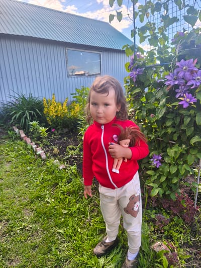A young child stands in a garden in Burayevo, Russia, holding a doll. The child is wearing a red hooded jacket and light-colored pants with a teddy bear graphic. Behind the child is a garden bed with various plants, including yellow flowers and purple clematis climbing a trellis. A light blue, corrugated metal building with a window is visible in the background. The sky is cloudy.