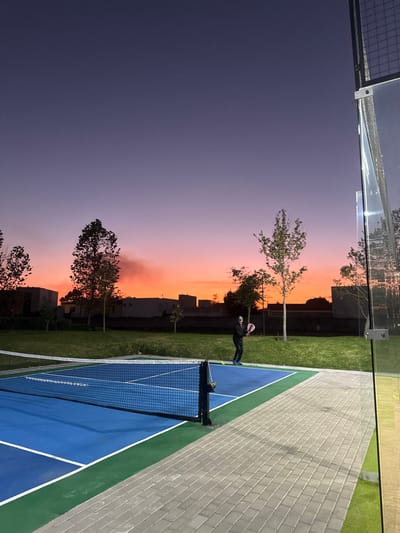 A tennis court, blue with white lines, sits beside a green lawn. A man dressed in dark attire plays tennis, holding a ball above his head. The background features buildings and trees silhouetted against a vibrant sunset sky, which transitions from orange near the horizon to deep purple overhead. A grey tiled walkway extends from the court.