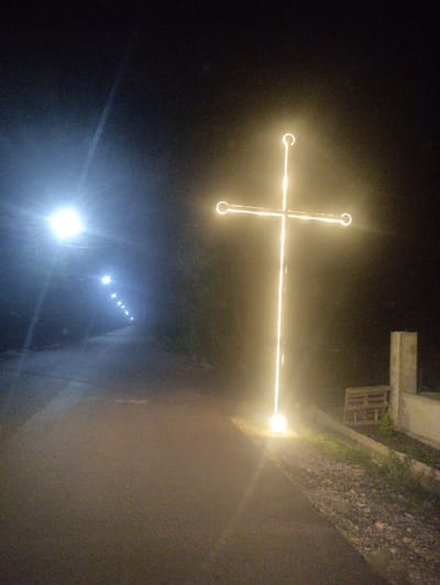 This media file shows a roadside scene at night in Tamarisi, Georgia. The primary subject is a large, illuminated cross standing prominently on the right side of the frame. The cross is constructed from glowing yellow light, possibly neon or LED, and is the most brightly lit object in the image.
To the left, a road curves away into the distance, lined with a series of streetlights that also emit a bright white light. The road surface is dark asphalt, and the area surrounding it is obscured by darkness and vegetation. In the far distance, the streetlights create a diminishing perspective, suggesting a long, straight stretch of road.
On the right side of the road, near the cross, there are some visible objects: a concrete structure, possibly a low wall or plinth, and a wooden pallet. The overall mood is serene and perhaps spiritual, given the prominent cross. The time of day is clearly night, with no indication of weather conditions other than the darkness. The location context is explicitly stated as Tamarisi, Georgia. There is no visible text in the image.