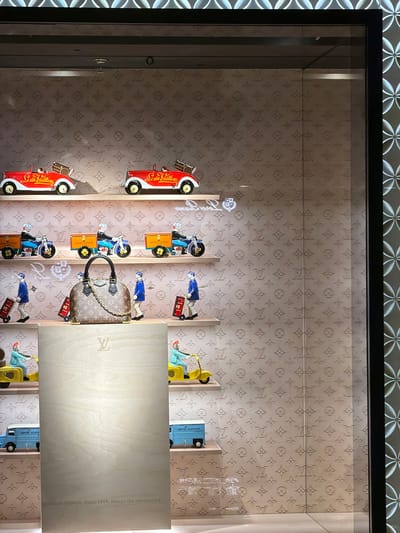 The image depicts a retail display window, likely within a Louis Vuitton store in Xuanwu District, China. The display is organized into multiple shelves. The background of each shelf features a repetitive pattern of the Louis Vuitton monogram.
The top shelf showcases two red toy cars with white accents, each carrying a miniature brown Louis Vuitton trunk on its rear. The second shelf from the top displays three toy motorcycles, each ridden by a miniature figure dressed in a blue uniform and cap, pulling a yellow cargo box.
The third shelf features a central, medium-sized Louis Vuitton handbag with a prominent gold chain strap. Flanking the handbag are four miniature figures dressed in blue uniforms and caps, each interacting with small, red rolling suitcases.
The fourth shelf from the top has two yellow toy scooters, each ridden by a miniature figure wearing a blue uniform and red hat.
The bottom shelf is dominated by a large, light-colored wooden panel, seemingly a display stand. A Louis Vuitton logo is embossed near the top of this panel, and text at the bottom reads "Louis Vuitton. Since 1854, always the unexpected." On either side of this wooden panel, at the very bottom of the visible display, are two light blue toy vans, each with "LOUIS VUITTON JOUETS" printed on its side.