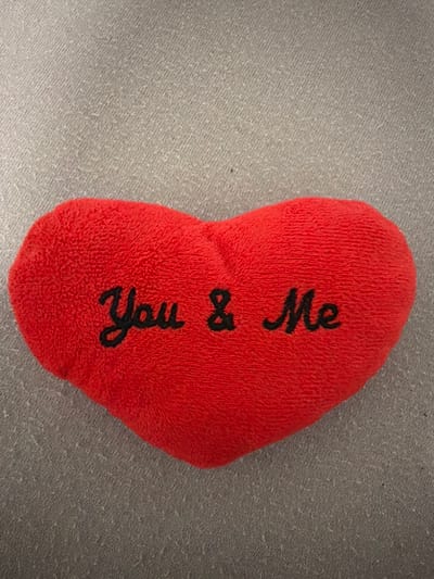 A red heart-shaped plush toy with black embroidered text that reads "You & Me" is lying on a textured gray fabric surface. The plush toy is slightly angled in the center of the frame. The gray fabric appears to be a tablecloth or similar textile with a woven pattern. There is no identifiable location within the city of Sankt Augustin, Germany.