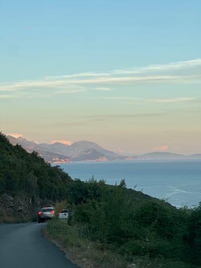 An eye-level shot captures a scenic view of the coast of Višnjevo, Montenegro, as two cars drive along a winding road. The road curves to the right, disappearing into the lush green foliage that covers the left side of the frame. The dense trees and bushes form a natural barrier against the rocky hillside. On the right side of the road, wild grass and smaller plants grow.

The first car, a light-colored SUV, is in the foreground, heading away from the viewer. Its brake lights are illuminated, suggesting it is slowing down. Behind it, a white car is also visible, further down the road, its brake lights also on.

The road leads to a vast expanse of the Adriatic Sea, which dominates the right half of the image. The water is a calm, deep blue, with gentle ripples on its surface. In the distance, a chain of mountains stretches across the horizon, their peaks softened by the atmospheric haze. A few scattered lights, possibly from a coastal town, are visible along the base of the mountains.

The sky above is a gradient of soft colors, transitioning from a pale blue at the top to a warmer, pastel hue near the horizon. Wispy clouds are scattered across the sky, catching the light of what appears to be a late afternoon or early evening sun. The overall impression is one of tranquility and natural beauty.