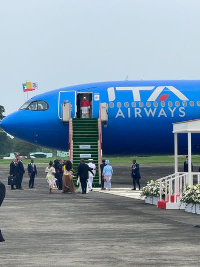 A large blue ITA Airways aircraft is parked on an airport tarmac in Malabo, Equatorial Guinea, serving as the setting for an official arrival or departure ceremony. The aircraft, with its name "ITA AIRWAYS" prominently displayed in white and red lettering along its fuselage, has green-carpeted airstairs extended from its forward door.

At the top of the airstairs, within the open aircraft doorway, a figure identifiable as the Pope, dressed in a white cassock and red cape, is visible, looking out at the gathering on the ground. On the aircraft's nose, two small flags are mounted: one displaying the Vatican colors (yellow and white), and another, likely representing Equatorial Guinea.

On the tarmac below, a group of people, including men in dark suits and women in various formal attire, are assembled. They appear to be dignitaries forming a receiving line or a farewell committee. In the mid-ground, a woman in a distinctive blue patterned dress stands out among the group. Further back, a ceremonial setup featuring white railings adorned with lush white and yellow floral arrangements adds to the formal atmosphere.

The scene takes place during daytime under a clear or lightly overcast sky. The overall impression is one of a formal and respectful official event at an airport.