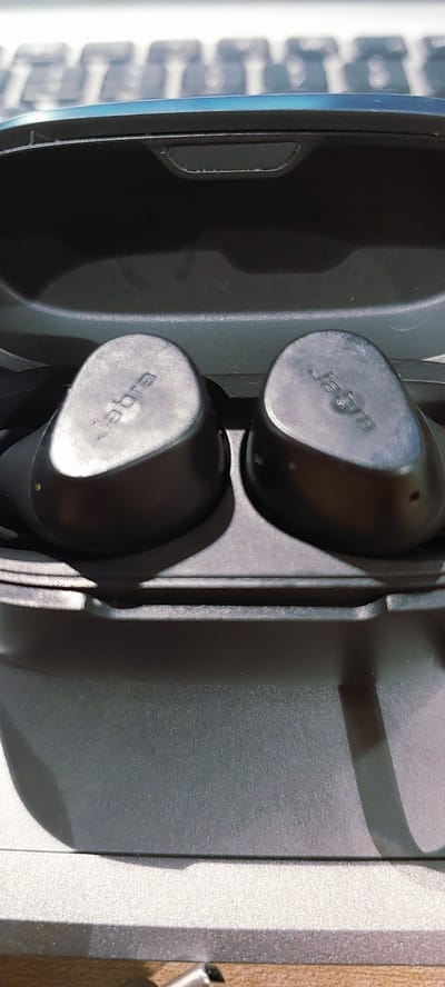 The image shows a pair of black Jabra wireless earbuds in their charging case. The earbuds are positioned side-by-side, with the Jabra logo visible on each. The charging case is dark gray and appears to be open, revealing the indentations where the earbuds sit. The background is out of focus, but suggests a keyboard-like surface with a bluish tint. The overall lighting is subdued, with shadows cast on the charging case. There are no people or animals present in the image, and no signs of the location in Pagedangan, Indonesia.