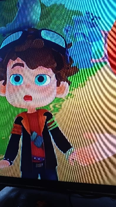 The scene depicts a digital photograph of a screen displaying an animated character. The character is a young, cartoon-style male figure with dark brown hair, light blue eyes, and an open mouth suggesting surprise or alarm. He wears black goggles pushed onto his forehead, an orange and red short-sleeved shirt with a blue geometric pattern on the chest, a black long-sleeved jacket with white horizontal stripes on the left sleeve, and dark pants. His hands are visible at his sides, with the left hand partially obscured. The animated background features abstract shapes and colors, predominantly green and blue on the left and yellow-orange on the right, displaying vertical and diagonal scan lines characteristic of a captured screen image. A dark, black frame of a television or monitor is visible at the bottom edge of the image, with an indistinct dark surface directly below it. The image originates from the city of Kasansay, Uzbekistan.