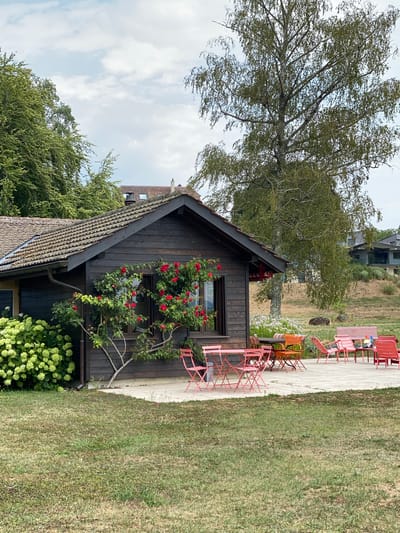 The image shows a wooden cabin with a dark brown exterior and a tiled roof, set in a grassy area. Red roses are climbing the side of the cabin near a window. In front of the cabin, there is a patio with several sets of pink metal outdoor furniture, including chairs and tables. To the left of the cabin, there is a large bush with green flowers. In the background, there are trees and other buildings on a hill. The sky is cloudy. The scene appears to be a peaceful outdoor setting, possibly a cafe or a private residence, located in Laténa, Switzerland. There are no people visible in the image.