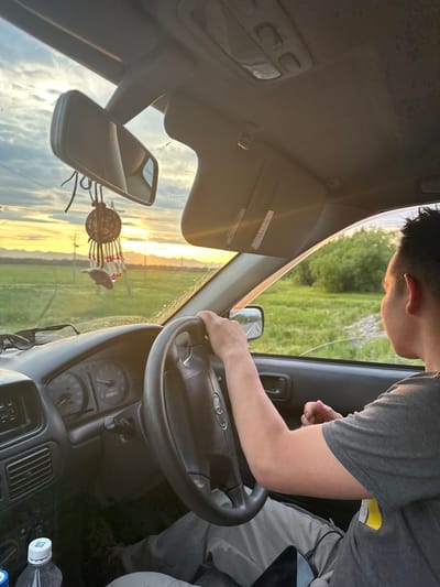 The image shows the interior of a car, with a person driving. The driver's right hand is on the steering wheel. Outside the car, there is a field of green grass under a cloudy sky with the sun setting behind distant hills. The scene is peaceful and idyllic.