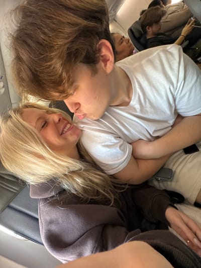 A young man and woman are seated in an airplane. He has brown hair and a white t-shirt, while she has blonde hair and wears a brown hooded sweatshirt. They are close together, heads almost touching, and she is smiling. In the background, other passengers can be seen seated. The setting appears to be during a flight, likely a commercial airline.