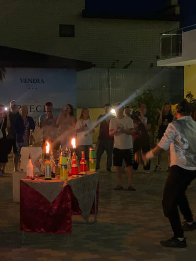 In the city of Vityazevo, Russia, a group of people are gathered outdoors at night, possibly at a resort as indicated by a sign reading "VENERA RESORT". The scene appears to be a performance or demonstration involving flaming bottles, likely a cocktail show. In the foreground, a table is set up with various bottles, some of which are lit and emitting flames. People stand around the table, observing the display, with some holding up phones, suggesting they are capturing the event. A person in the mid-ground, possibly the performer, is visible, and a spotlight beam cuts across the scene from the left, illuminating the group. The environment is paved with interlocking stones, and a brick building is visible in the background.