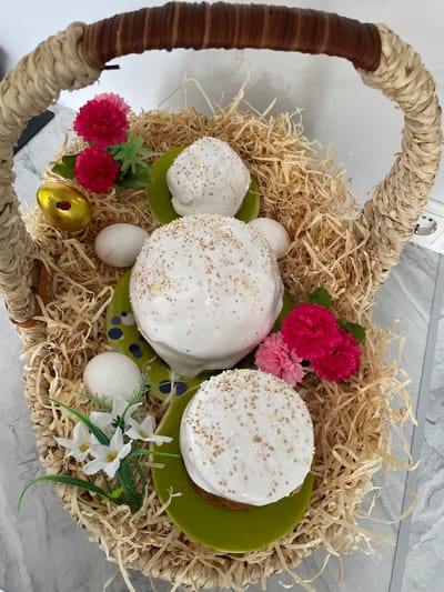 The image shows a wicker basket filled with straw-like material. Inside the basket are three decorated cakes, likely Easter cakes, topped with white icing and sprinkles. They sit on green plates. Also within the basket are three white eggs and one golden egg. Floral decorations, including pink and white flowers with green leaves, are scattered around the cakes and eggs. The handle of the basket is visible on the left and top of the image. The background is a light gray surface, possibly a countertop. The location of the image is Лесной Пейзаж Кп (шарапово, Russia.