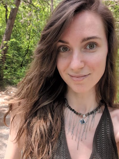 A young woman with long, brown, wavy hair and light green eyes smiles gently at the camera. She is wearing a dark, crocheted halter top and a beaded necklace with silver chain accents and a clover pendant. She is outdoors, with a blurred background of trees and foliage, suggesting a natural, possibly forested, setting. The ground appears to be a dirt path. The overall lighting is natural and bright, indicating daytime. The location is not specified to be in Dzhankhot, Russia.