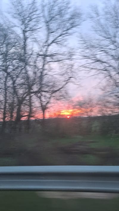 The image shows a blurred sunset through a window. Bare tree branches are visible in the foreground against the sky. A red glow is seen on the horizon, likely indicating the setting sun. A dark green field is present to the right. The edge of the window frame is visible on the right side of the image. The image is taken from inside, looking outward. No people are visible. The location in Donchevo, Bulgaria is not confirmed in the image.