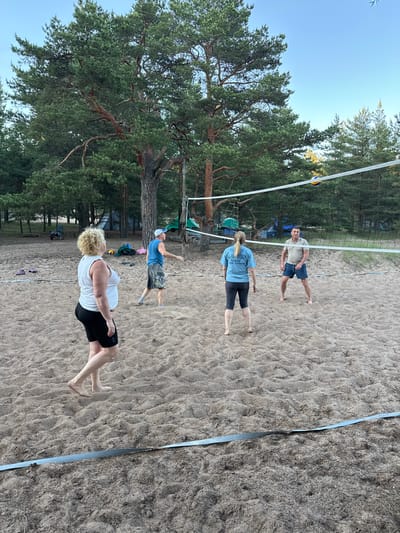 Four people are playing volleyball on a sandy court in Ozerki (Krasnodolinskaya), Russia. The court is situated amidst a pine forest, with tents and camping equipment visible in the background. The sky is clear and blue. The individuals are casually dressed, with two women and two men on the court. One woman, with blonde curly hair, is wearing a white tank top and black shorts. Another woman, with blonde hair tied back, is wearing a light blue t-shirt and dark leggings. A man in a blue tank top and patterned shorts is standing near the net. The fourth person, a man wearing a light brown polo shirt and blue shorts, is on the opposite side of the net. A volleyball is visible in the air above the net. The sand on the court is unevenly distributed, with footprints and debris scattered across it. The trees surrounding the court are tall and dense with green pine needles.