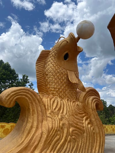 The image depicts a large, ornate golden statue of a fish, positioned outdoors in Đăk Mar, Vietnam, beneath a clear blue sky dotted with scattered white cumulus clouds. The statue, rendered in a textured golden material, features distinct scales and fins. Its head is angled upwards with an open mouth, which holds a smooth, pearlescent white spherical object. A dark, hollow eye socket is visible on the fish's head. The lower portion of the statue is sculpted into stylized, swirling golden waves. In the background, dense green foliage and trees are visible on both the left and right sides of the frame. A low golden wall or barrier, adorned with yellow relief carvings resembling lotus flowers, is partially visible at the bottom left and far right. No individuals are present in the scene.