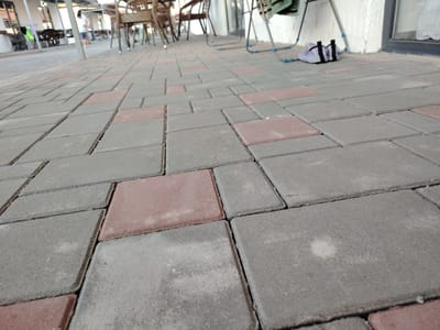 The image depicts an outdoor paved area with interlocking concrete pavers, some of which are reddish-brown, and others are gray. The pavers are laid in a staggered pattern. In the background, there are tables and chairs, suggesting an outdoor seating area, possibly for a cafe or restaurant. A person in a white t-shirt and blue shorts is visible in the distance, walking. There are also some metal chairs and a white wall in the background. To the right, a pair of white sneakers with black straps is placed on the pavement, near the wall. The scene appears to be in a public outdoor space, likely in the city of Toranghylyq, Kazakhstan.