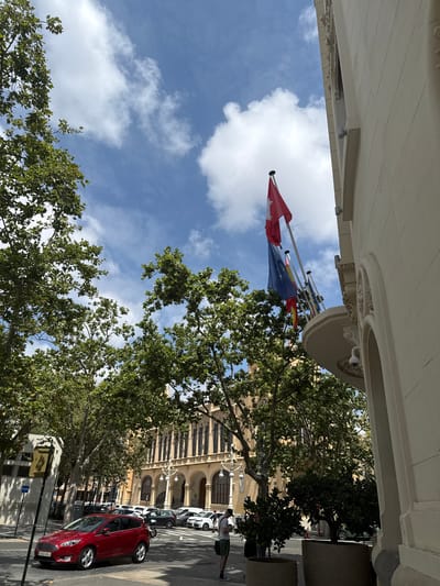 This media file depicts a street scene in Alboraia, Spain, likely during daytime given the bright sunlight casting shadows. The perspective is from beneath an overhang of a building, looking upwards and outwards.
The primary subjects include several flags flying from poles, a large green tree with dense foliage, and a street lined with parked cars. The flags appear to be the European Union flag and the Spanish flag, along with potentially others. In the background, a multi-story building with arched windows and architectural details is visible. Parked on the street are several cars, most notably a bright red hatchback in the foreground.
While no people are clearly in focus or prominently featured, there are glimpses of individuals, possibly pedestrians or people in the background, blurred or partially obscured by the foliage and distance. The activity is static, focusing on the setting and its elements.
Notable details include the architecture of the building, suggesting a potentially historical or significant structure. The weather appears to be clear and sunny, with scattered white clouds in the blue sky. The presence of parked cars and pedestrian crossings implies an urban or semi-urban environment.
Visible text includes a license plate on the red car which reads "5583 HWJ". There is also a blue sign with "TAXI" written on it. The overall mood is one of a typical sunny day in a European town.