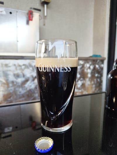 The image depicts a filled Guinness glass with a bottle of Guinness next to it. The background includes a countertop and an out-of-focus area showing a possible service area. The location cannot be determined to be Schmelz, Germany with the information provided.