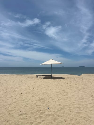 An umbrella and a lounge chair sit on a sandy beach in Hoi An, Vietnam. The umbrella is white and casts a shadow on the sand. The lounge chair is made of wood and has a metal frame. The beach is empty except for the umbrella and chair. The ocean is calm and blue, and the sky is clear with a few scattered clouds. In the distance, there are a few small islands.
