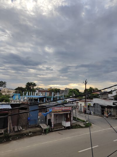 The image shows a street scene in Sitarganj, India, under a cloudy sky. A paved road runs across the foreground, with a sidewalk or embankment bordering it. Several small shops with corrugated metal roofs and shutters are visible along the street. Some shops have awnings and signage, including one with the text "Sona Foot Wear" in Hindi. Electrical wires and poles are prominent features throughout the scene, crossing overhead and along the street. In the background, there are buildings, trees, and a distant horizon with hints of sunlight breaking through the clouds. The overall impression is a functional, somewhat weathered, commercial area on a day with overcast weather. There are no people visibly present in the scene.