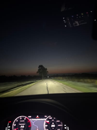 The image captures a first-person perspective from inside a vehicle at dusk or early night, looking forward through the windshield onto a rural road in Blienschwiller, France. The sky transitions from dark blue overhead to an orange-red glow near the horizon, with a single bright celestial body visible in the upper-left sky. The two-lane asphalt road ahead is illuminated by the vehicle's headlights, revealing cracks in its surface. Green grassy verges line both sides of the road, blurring into darker, distant vegetation and a treeline that defines the horizon. A prominent, light-barked tree stands on the left side of the road in the mid-distance, casting a long shadow across the pavement. The bottom portion of the image shows the vehicle's digital instrument cluster, displaying a speedometer indicating 80 km/h, an RPM gauge with a reading of 11.6, and a navigation map. The navigation system partially displays "Bliens," corroborating the Blienschwiller location. A reflection of an infotainment screen, showing various application icons and a music player interface, is visible in the upper-right area of the windshield. No individuals are present in the scene.
