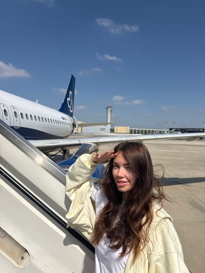 A young woman with long brown hair stands on the steps of an airplane, waving her hand towards the camera. She wears a white t-shirt and a light yellow jacket. An airplane with a blue and white livery is visible behind her. The tail of the plane has a stylized logo. The background shows a bright blue sky with scattered clouds, the airport tarmac, and a control tower. The location is likely an airport in Yagel, Israel, based on potential contextual information.