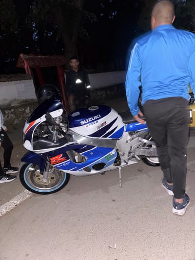 In Mihailovo, Bulgaria, a blue and white Suzuki GSX-R sport motorcycle is parked on a paved surface. A bald man wearing a bright blue athletic jacket and dark pants is walking away from the motorcycle, towards the right side of the frame. Another person, partially visible on the left, wears dark clothing and sneakers. In the background, a man in a dark hooded jacket stands near a red-roofed structure and a tree. The scene appears to be outdoors at night.