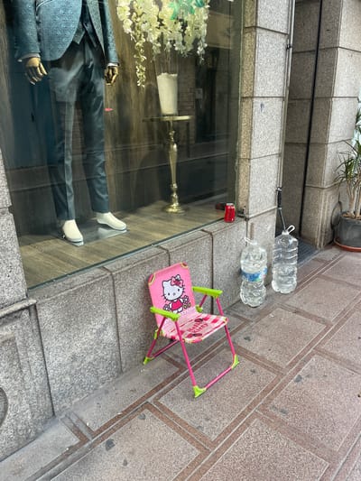 The image shows a street scene in La Línea De La Concepción, Spain. In the foreground, a child's pink folding chair with a Hello Kitty design sits on a sidewalk. Behind the chair, two large plastic bottles of water stand upright. To the left of the bottles, a red Coca-Cola can rests on a low stone ledge. Beyond the ledge, a shop window displays a mannequin dressed in a patterned teal suit, standing on a wooden floor. The building exterior is constructed of grey granite blocks.