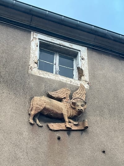 A building exterior features a weathered window with a flaking, white frame. To the right of the window is a stone sculpture of a winged lion-like creature holding a scroll. The facade has a rough, gray texture, and a dark gutter runs along the roofline visible in the upper left corner. Three bolt heads are apparent near the figure.