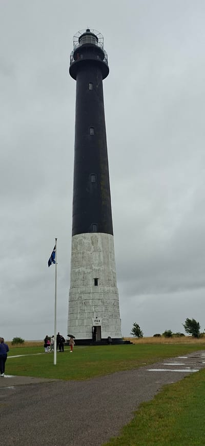 A tall, tapering lighthouse with a black top section and a white, weathered lower section stands against a cloudy sky. The lighthouse has several small, rectangular windows and a lantern room at its peak. A flag pole with a blue and white flag is positioned to the left of the lighthouse. Several people, some holding umbrellas, are gathered near the base of the lighthouse. The ground in front is covered with green grass and a paved path. The lighthouse is labeled with the year "1960". The scene is the Sääre lighthouse on the Sõrve Peninsula in Estonia.