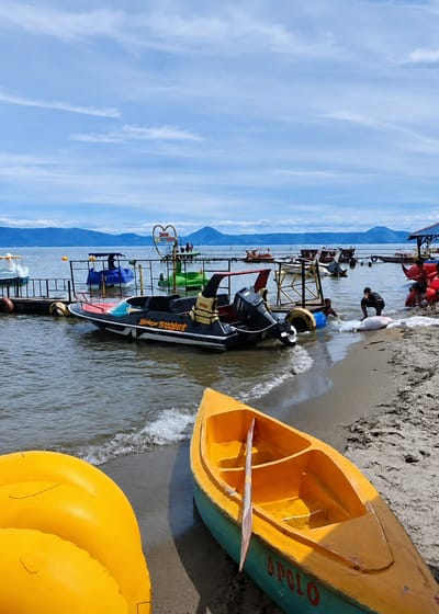 A scenic view captures the shoreline of Situngkir, Indonesia, with the tranquil waters of a lake meeting a sandy beach under a vast, partly cloudy sky. Several colorful boats, including a black and red jet ski labeled "Black Sweet," a blue pedal boat, and a bright yellow canoe with a paddle, are docked or resting on the sand. In the distance, mountains rise majestically against the horizon, adding to the picturesque landscape. A few individuals are present, with two men near the water's edge engaged in some activity, while others are visible on the pier. The overall atmosphere is serene and inviting, suggesting a popular spot for recreational water activities.