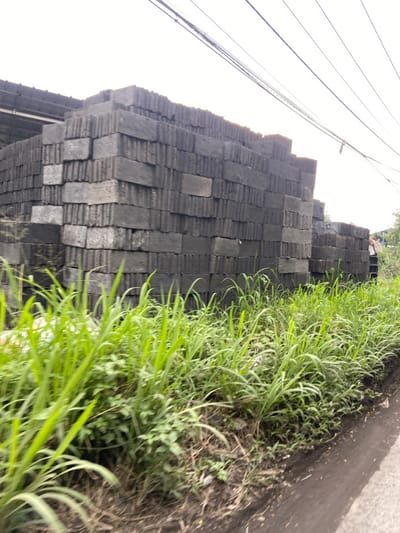 A stack of gray concrete blocks are piled high against a grassy embankment. Green grass and weeds grow along the embankment. The blocks are stacked neatly in a row and there are several power lines running overhead. The scene is located in the Kecamatan Margaasih region of Indonesia, at latitude -6.961 and longitude 107.536.