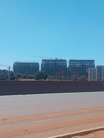 This image depicts a daytime scene in Gitaru ward, Kenya, dominated by urban development. In the foreground, a multi-lane asphalt road with white lane markings and a reddish-brown dirt shoulder is visible. A dark grey concrete barrier or retaining wall spans horizontally across the mid-ground, separating the road from the elevated terrain behind it.
Behind the barrier, several green trees and sparse vegetation are present. Utility poles carrying multiple lines stand on the left and center-right of this elevated area. A light-colored object, possibly discarded material, lies on the ground directly behind the barrier in the center-left. Two individuals are faintly visible behind the barrier: one in light clothing near a utility pole on the left, and another further right near the trees. Their specific actions are not discernible.
In the background, under a clear blue sky, multiple high-rise buildings are under construction. Three prominent structures show exposed concrete frames and extensive scaffolding, indicating ongoing development. A large construction crane is positioned between the two leftmost construction sites. To the far right, a completed or nearly completed multi-story apartment building with numerous windows and balconies stands, distinct from the active construction sites.