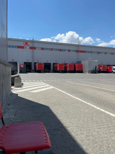 A wide, sunlit shot captures the exterior of a modern logistics facility on a clear day. The building, predominantly grey with a striking red horizontal band, features a long row of loading docks. Several red delivery trucks are parked at these docks, some with their doors open. Overhead, a sign with red lettering reads "2 УКРІТТЯ" (2 SHELTER), indicating a designated safety area. A metal ladder extends down the side of the building near this sign.

In the foreground, a grey, hexagonal-patterned paved area forms a parking lot or service yard. White painted lines mark pedestrian crossings and parking bays. To the left, a concrete wall and a portion of a red plastic chair are visible. A cylindrical concrete trash receptacle stands near the building entrance. The sky is a bright blue, dotted with fluffy white clouds. The scene appears to be a typical operational day at a postal or delivery service hub located in Nerubais'ke, Ukraine.