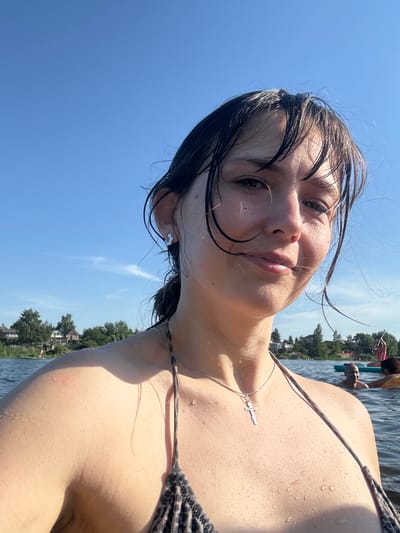 A young woman with wet, dark hair and water droplets on her skin is taking a selfie in a lake. She is wearing a leopard-print bikini top and a silver cross necklace. The background shows a clear blue sky with some wispy clouds, trees, and houses along the shore, suggesting a suburban or rural setting. Other people can be seen in the water in the background, some on paddleboards or inflatable rings. The image appears to be taken during the daytime, with the sun shining brightly. The location is identified as Pribrezhnyi, Russia.