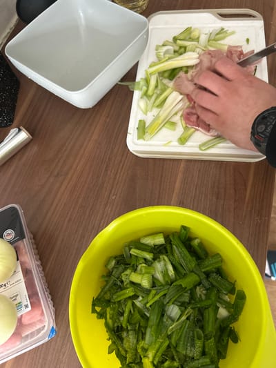 The image depicts a preparation scene of food. The individual's hand with a wristwatch on their wrist can be seen cutting meat with a knife on a white cutting board. Next to the meat is chopped leek. A yellow bowl filled with chopped green onions is nearby. A white square-shaped container sits to the left, presumably for storing the chopped items. At the bottom right of the frame, there's a packaged raw chicken and white onions.

The packaging reads "Fleischerei-Deutschland.de". Text on the packaging is in German, suggesting the location is in Germany. The packaging also contains the address of a "Platz" in Rüsselsheim am Main, Germany.